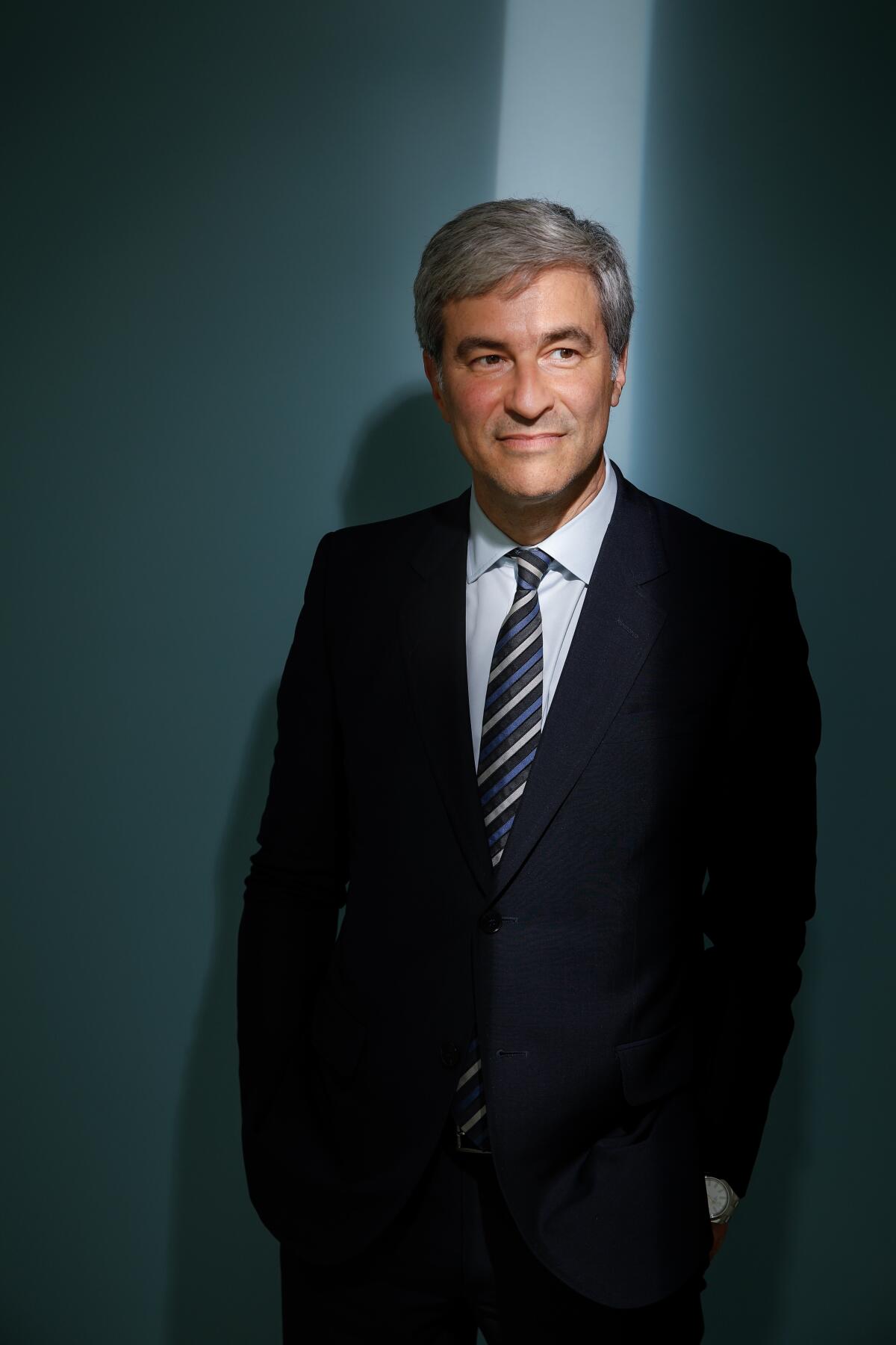 Michael Govan, with gray hair, wearing a dark suit and tie, stands in front of a blue backdrop and looks to the side. 