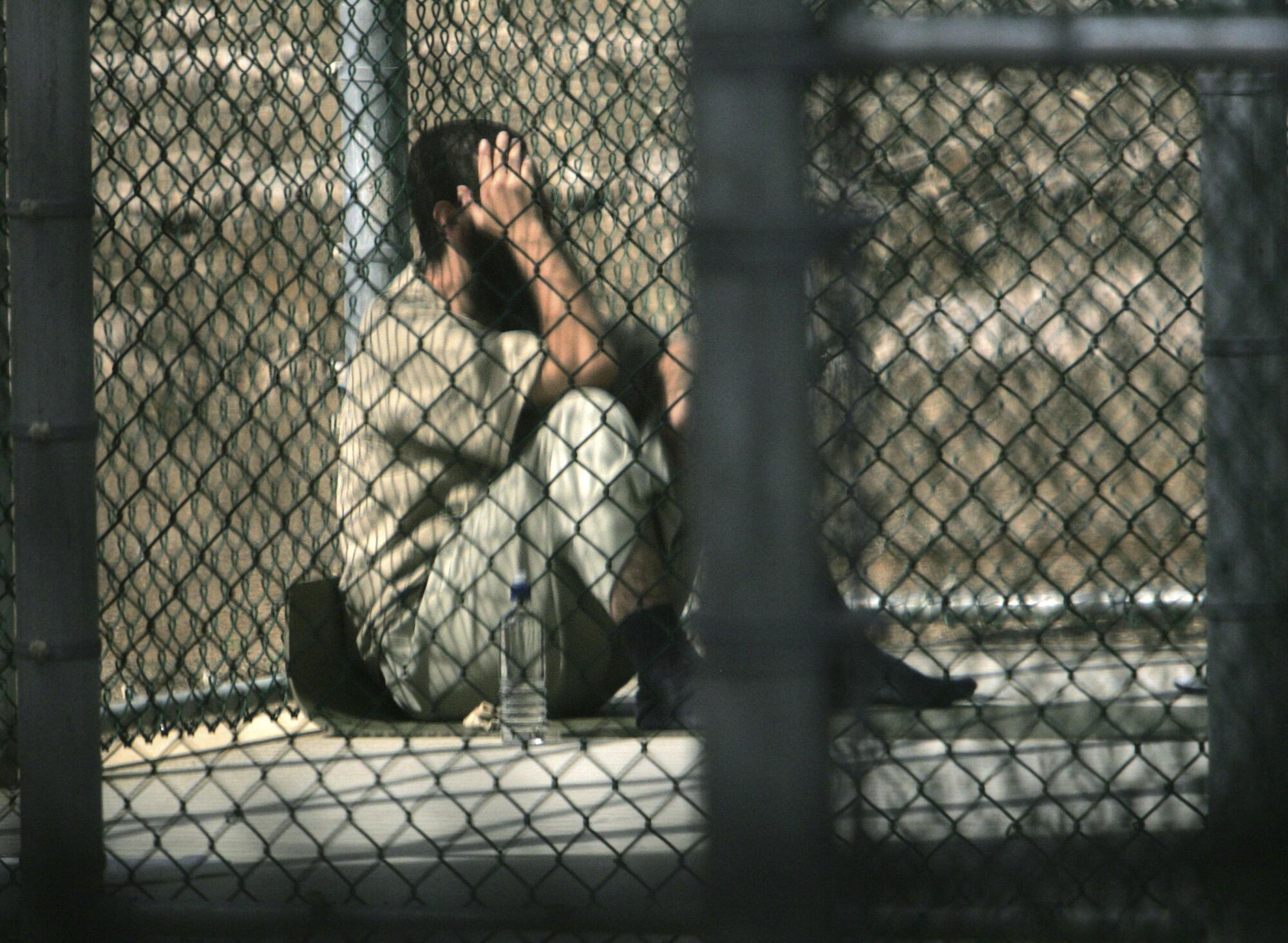 A man with dark hair and beard holds a hand to his face as he sits behind a wire fence 
