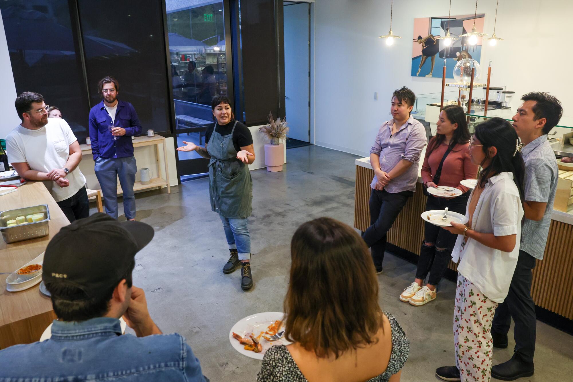 A woman in an apron stands in a kitchen, with other people standing around and sampling food