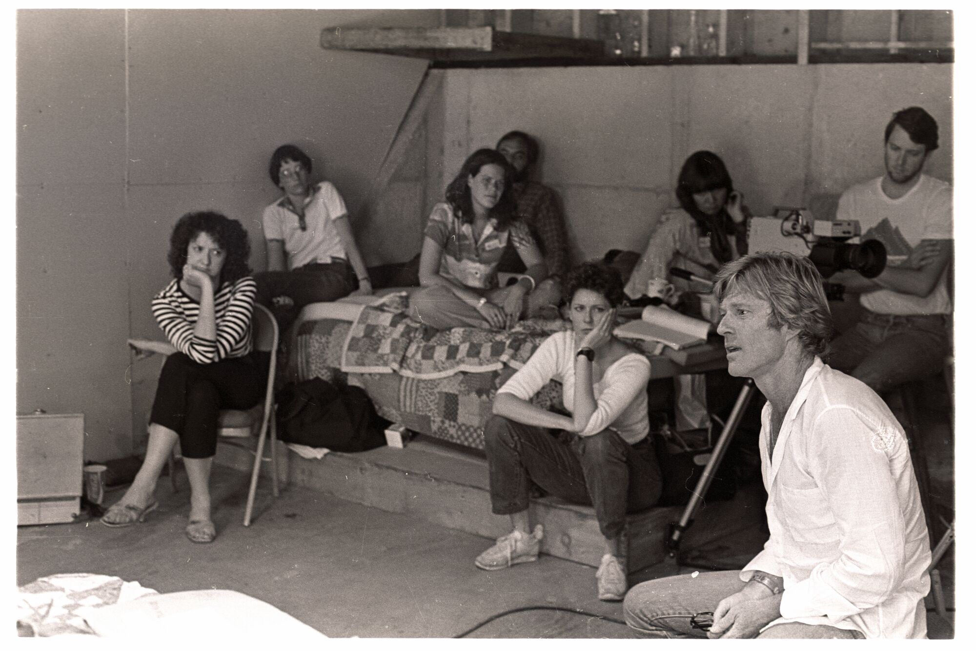 A black-and-white photo of seven people listening to one man, some on a bed