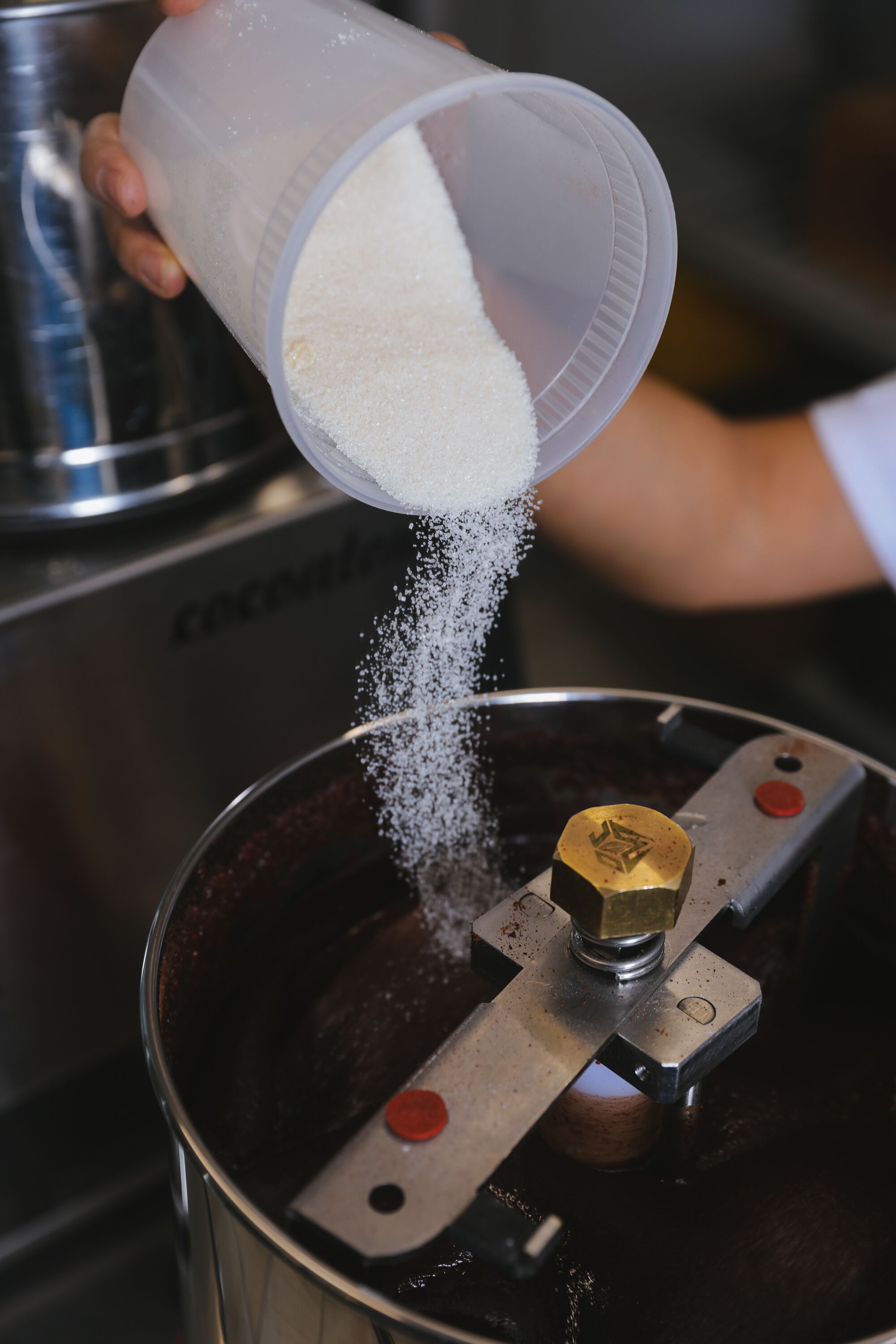 A hand pours sugar from a container into a mixing machine.