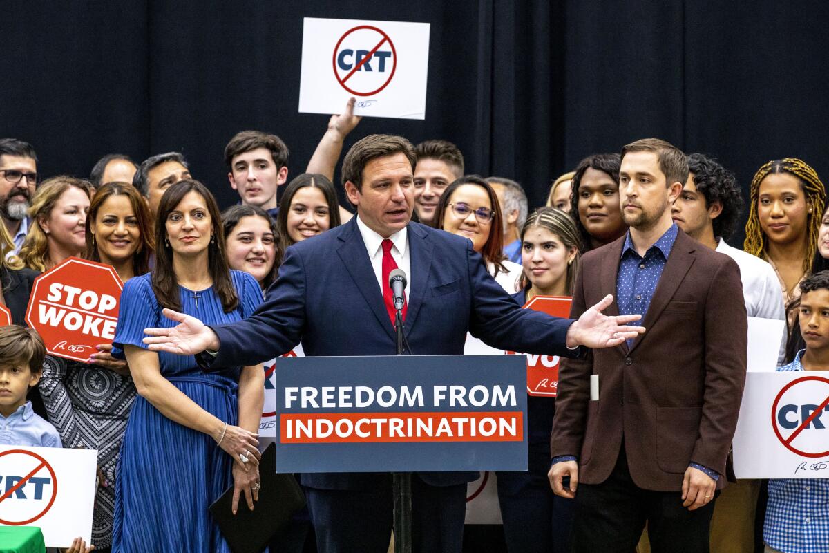 A man and supporters at a podium, with signs reading “Freedom From Indoctrination” and  “Stop Woke.”