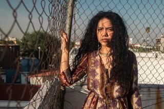 A woman with long dark hair stands next to a chain-link fence on a roof.