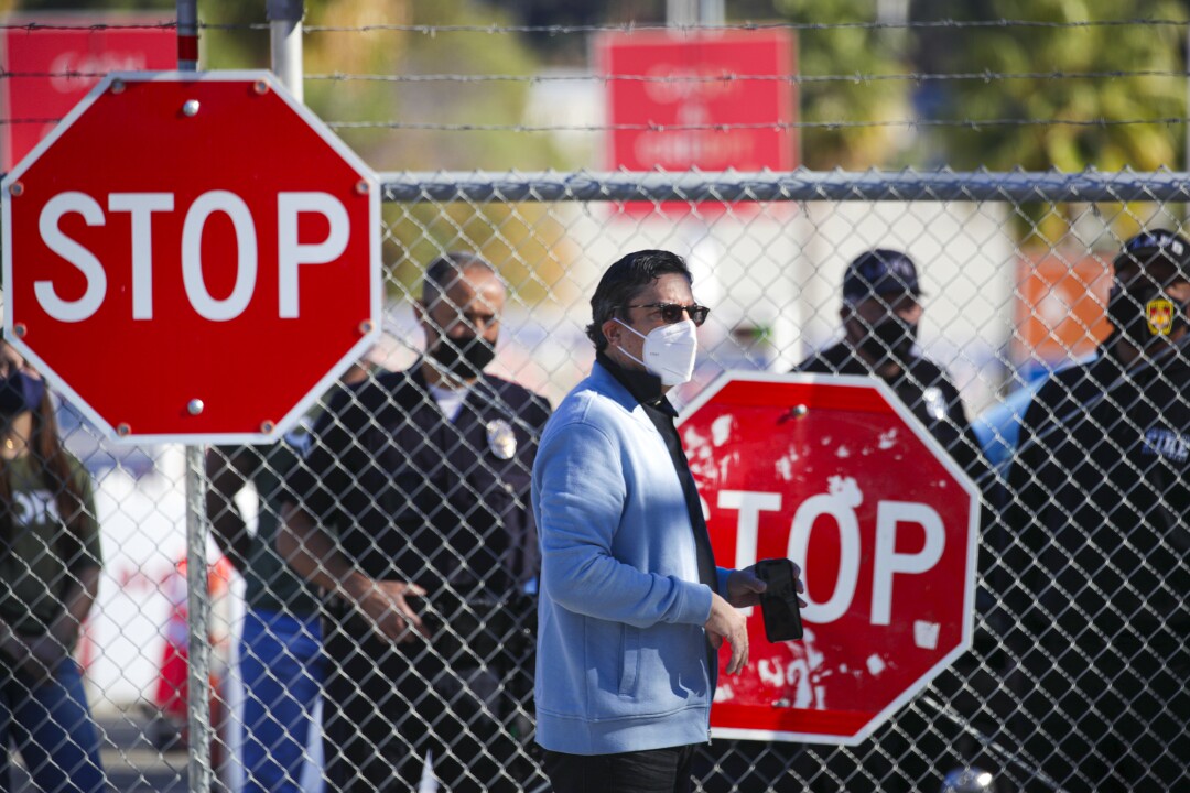 A man in a mask stands in front of a chain link fence, LAPD officers on the other side.