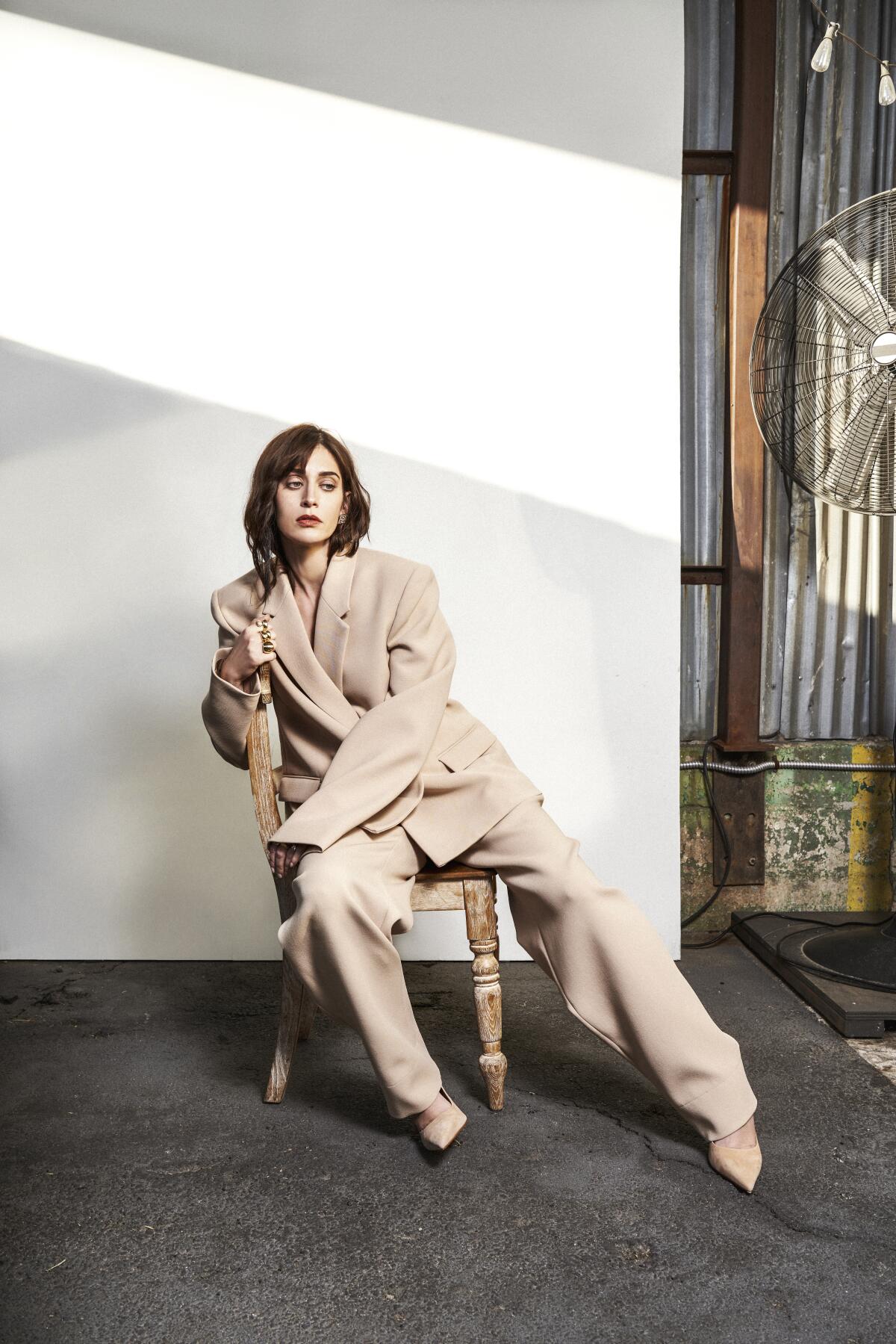 Lizzy Caplan, in an oversized suit, poses askew on a wooden chair. 