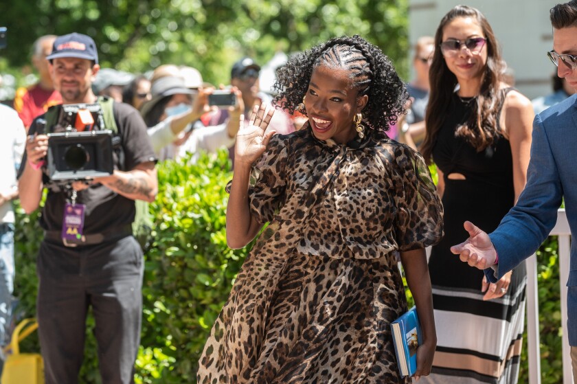 A young woman in a leopard print dress waves.  Around her, fans and photographers.