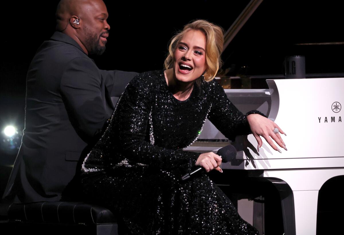 Adele wears a black beaded gown and leans over a white piano seat as a man plays the piano behind her
