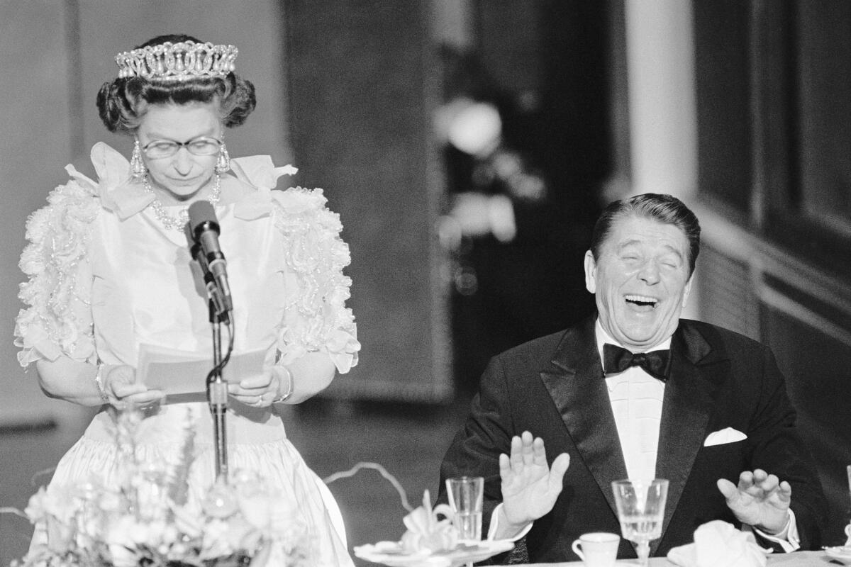 A woman in a tiara speaks into a microphone next to a man in a tux leaning his head back and laughing.