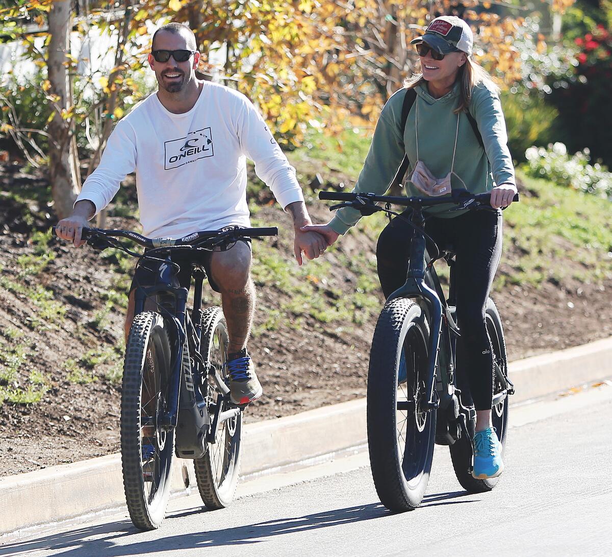 A man in glasses and a woman in glasses and a baseball hat hold hands while riding separate bicycles on a fall day.