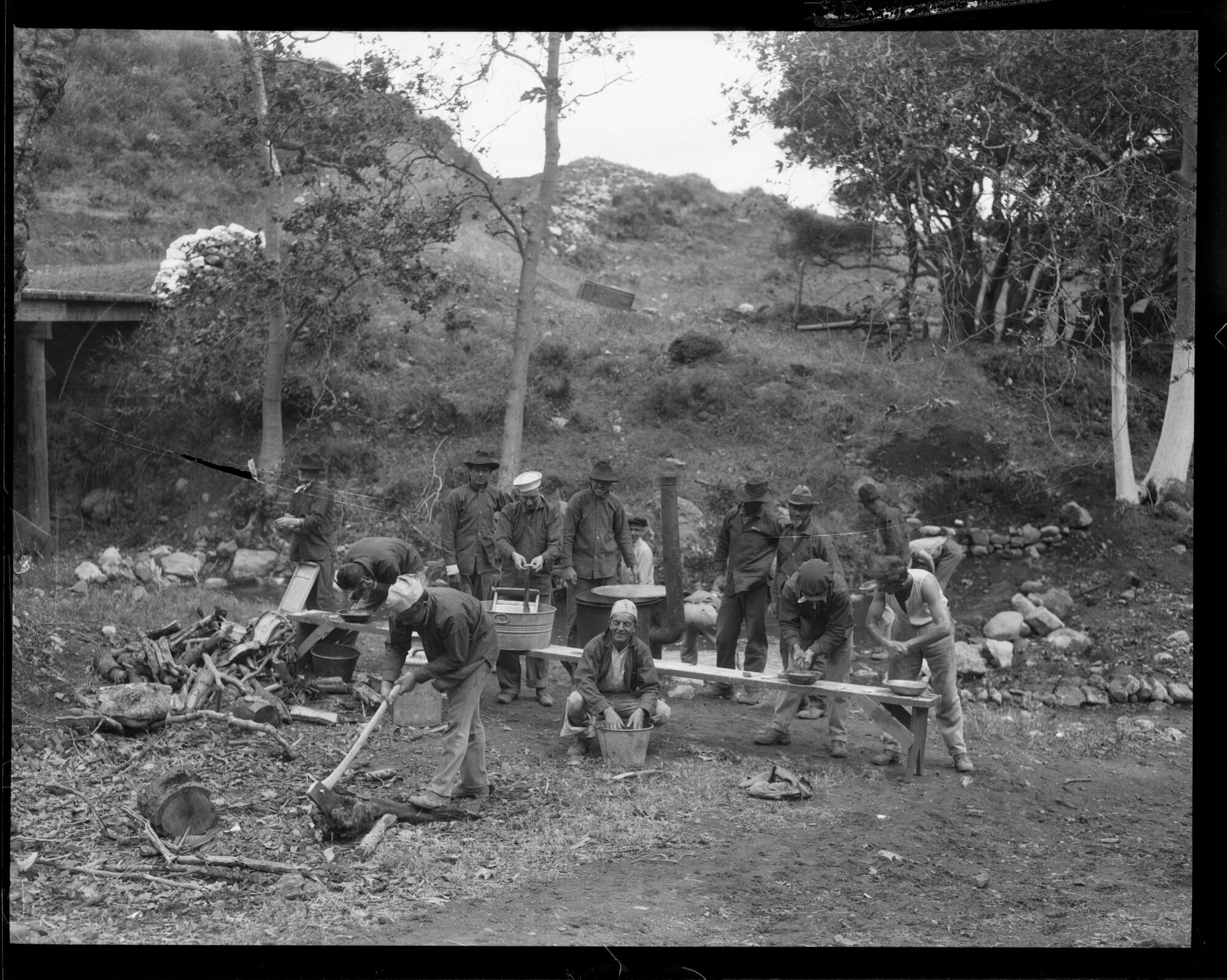 An open-air labor camp where men are seen washing themselves and their clothes with one man in the foreground chopping wood.