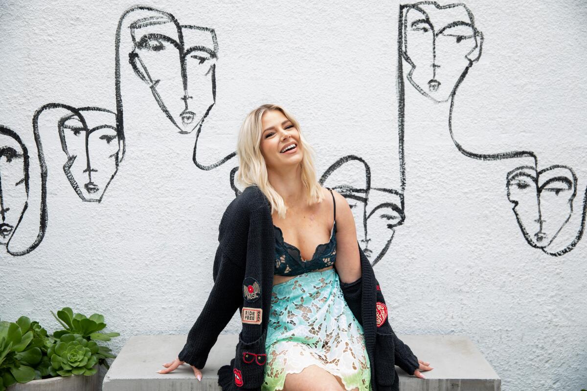 Ariana Madix sits in front of a restaurant mural with sketched faces. 