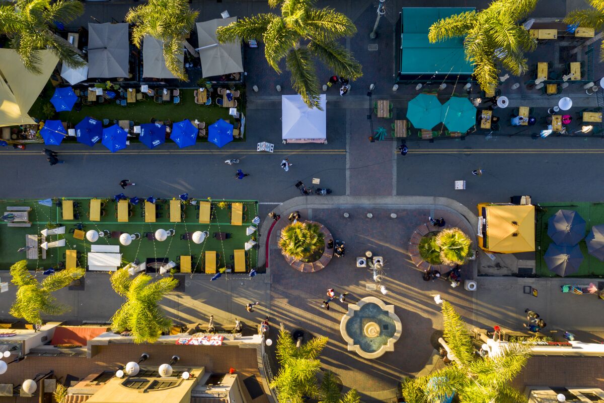 An overhead aerial view shows people walking, outdoor restaurant dining tables, umbrellas and a fountain.