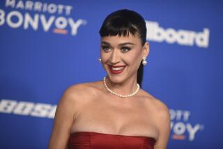 Katy Perry with short bangs posing in a pearl necklace and a strapless red gown against a blue background with white text