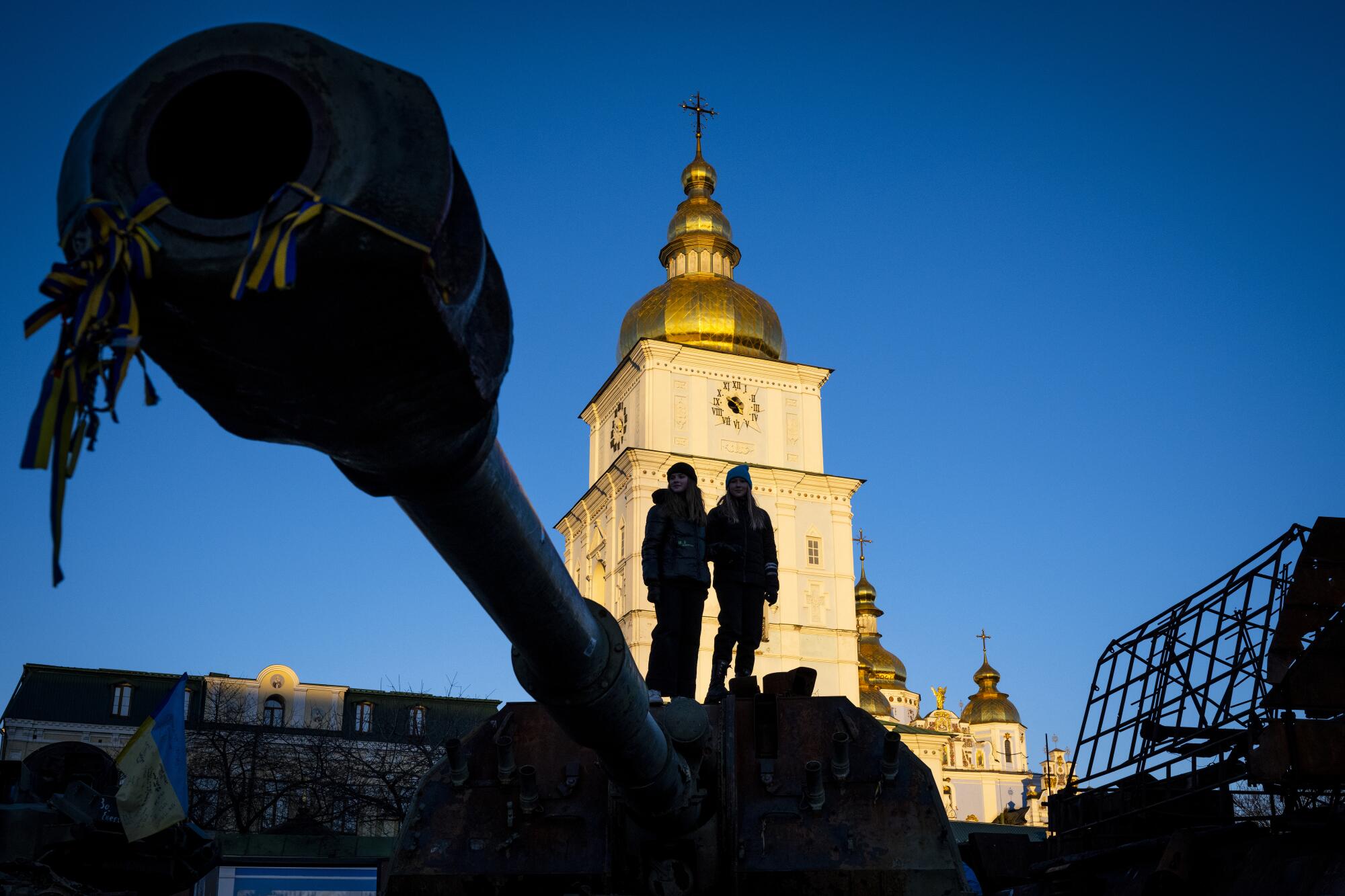 People view captured Russian equipment, including tanks, in front of a  golden-domed building.