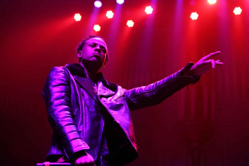 Rich Homie Quan in a leather jacket holding up his left hand as he performs on a stage under purple lights