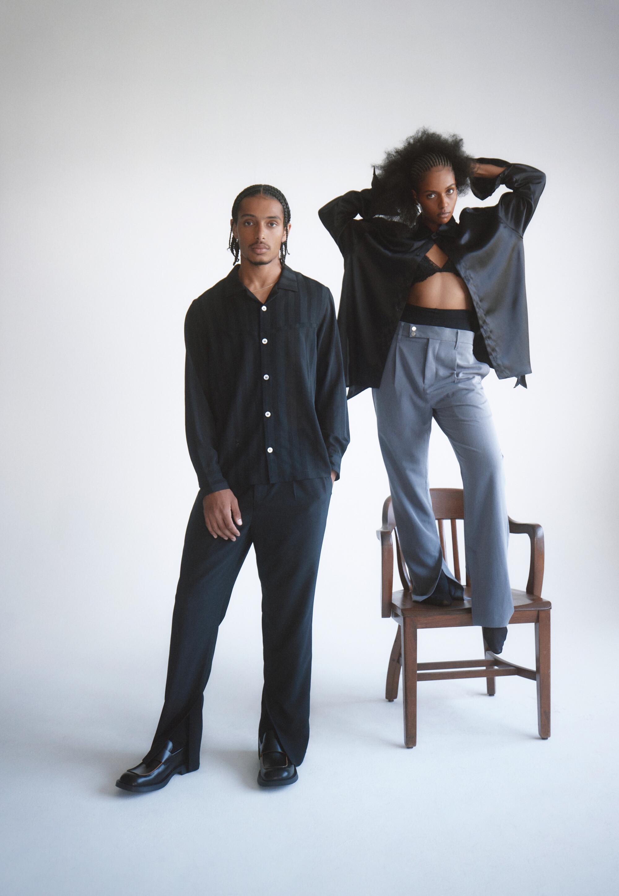 Standing to the left, a male model wears an all-black outfit. To the right, a female model poses, standing on a chair.