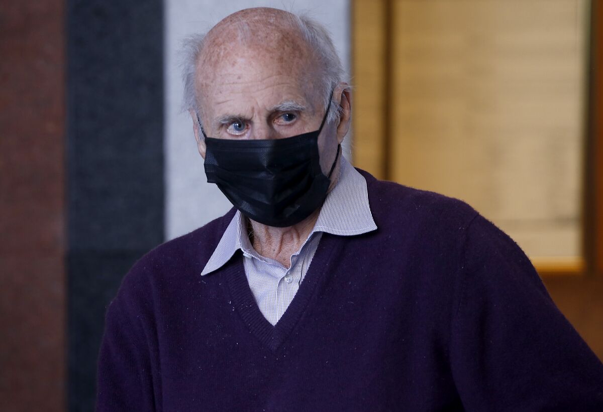 A head-and-shoulder image of an elderly-looking man in black facial mask, purple v-neck sweater and a collar shirt.