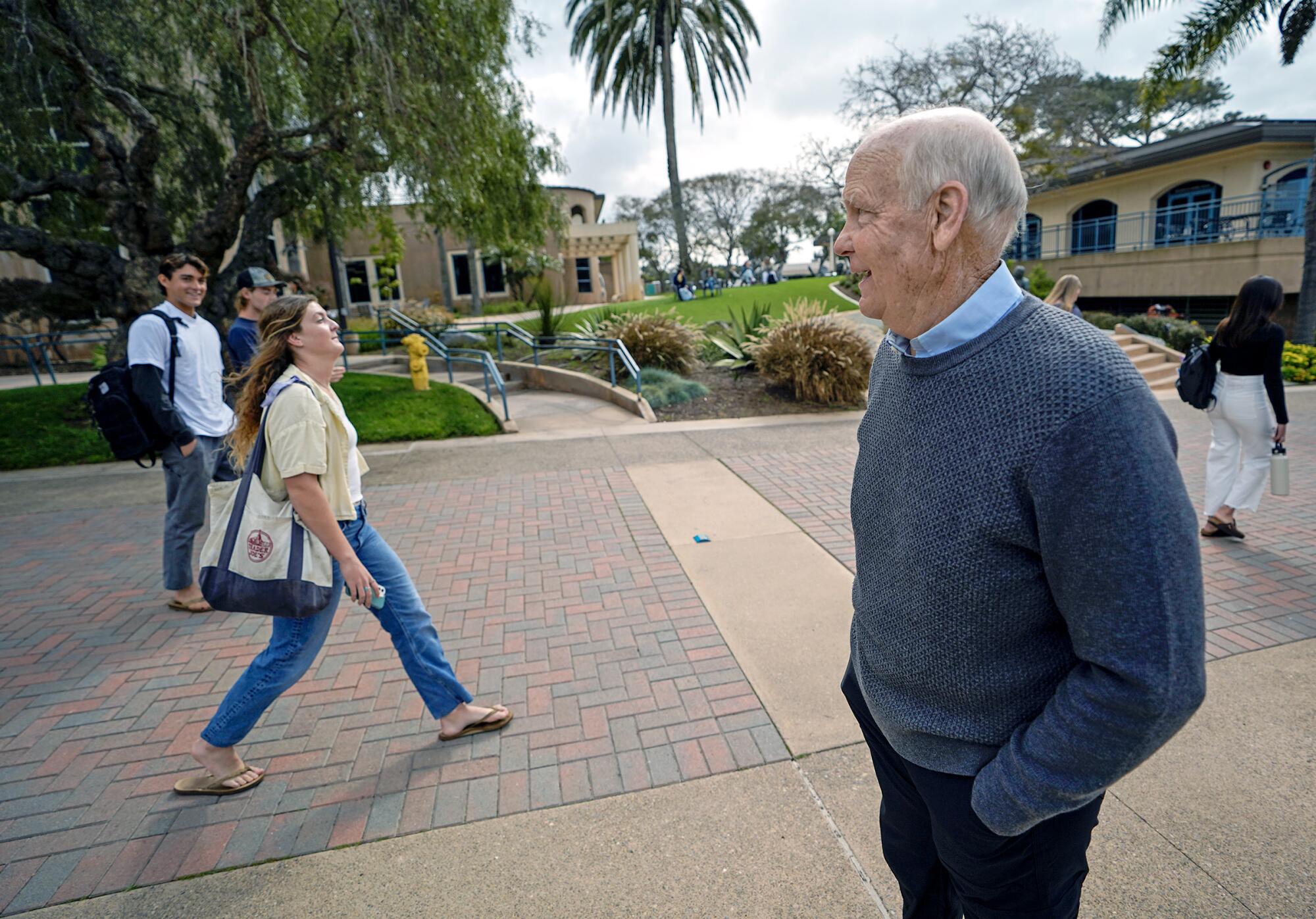 An older man in a sweater smiles as he greets college students on a campus walkway.