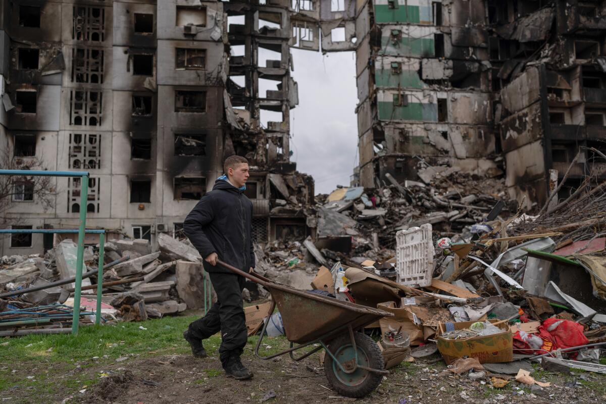 A young man pushes a wheelbarrow in front of a destroyed apartment building.