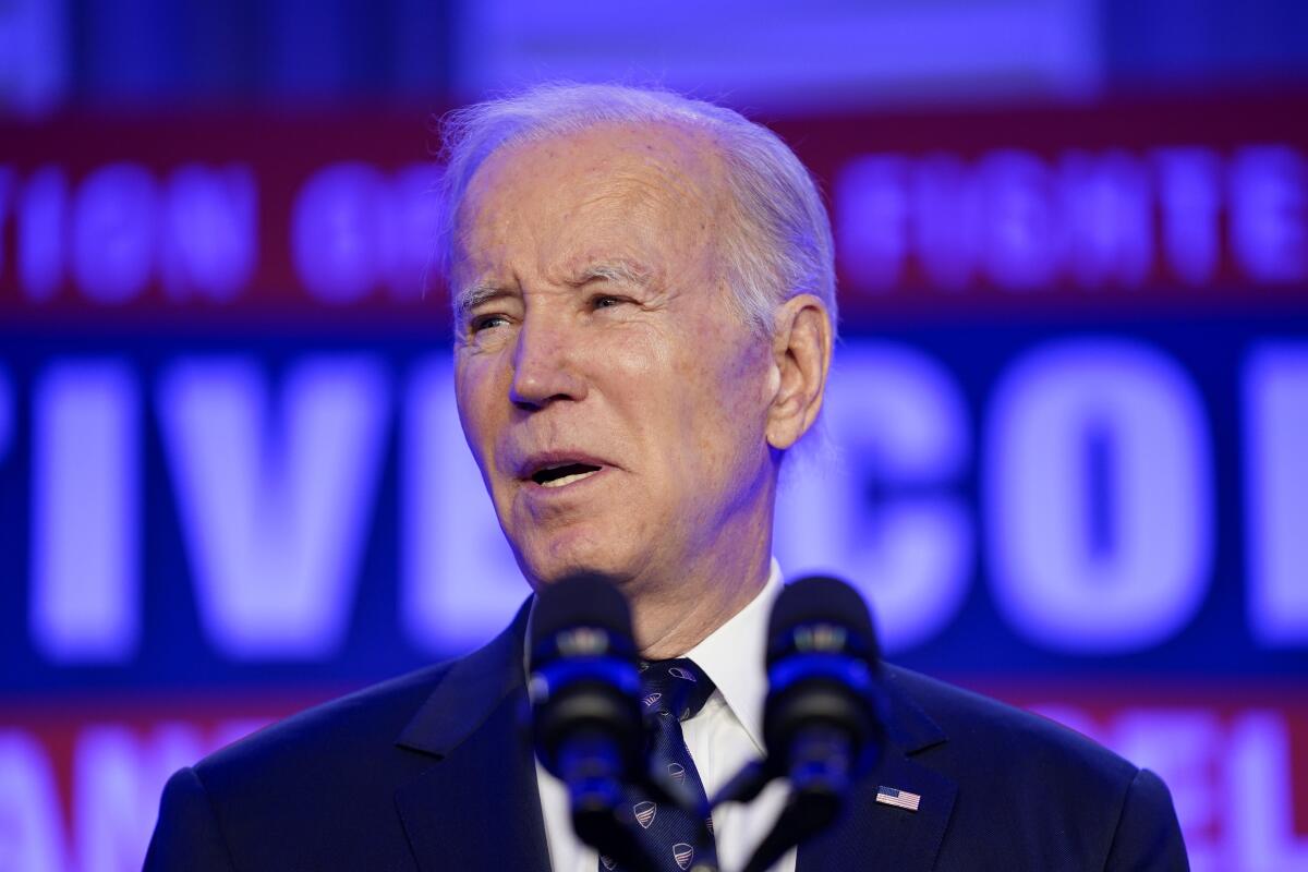 President Biden pictured from the shoulders up speaking into two microphones, against a blurry red, white and blue background