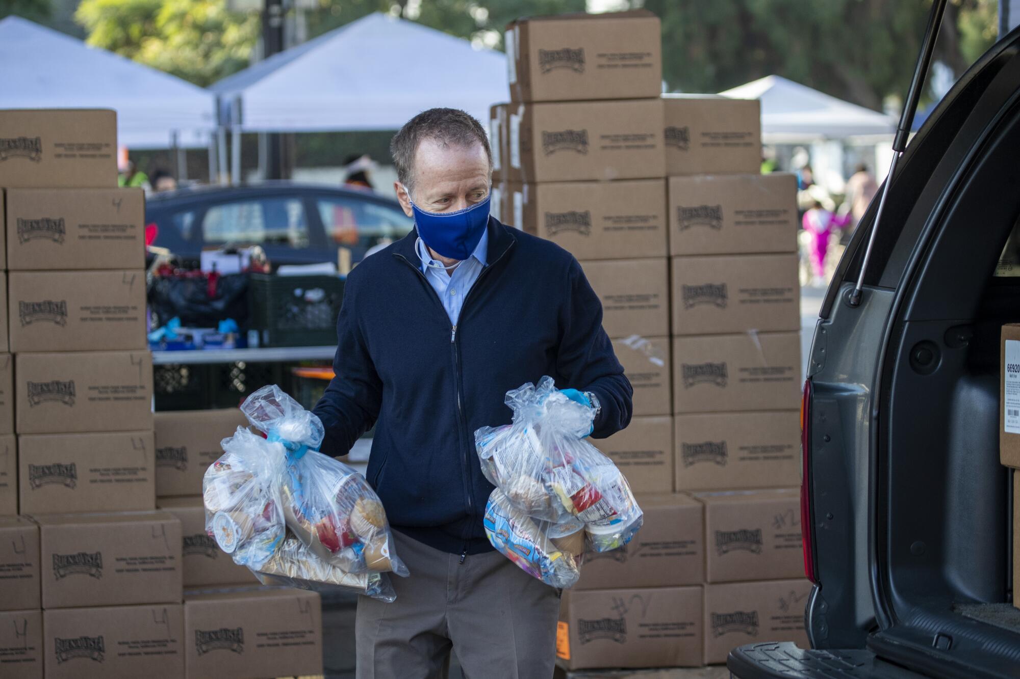 Superintendent Austin Beutner in a mask holds bags of food in each hand to load into a car