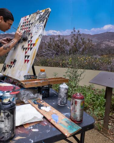 An artist paints outdoors, looking out at mountains and blue sky