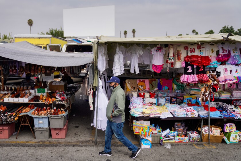 A man walks past wares, including food and children's clothing, beneath canopies in a parking lot.