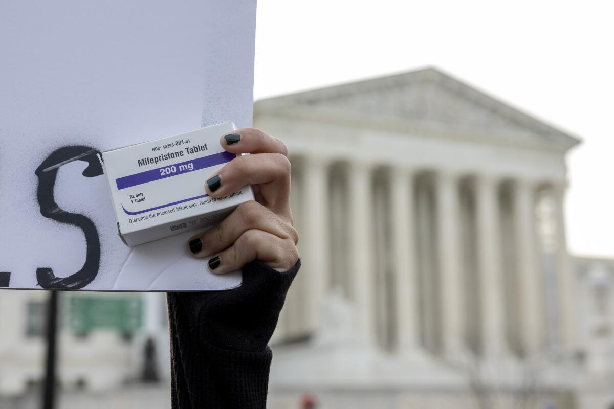 A woman's hand with black nail polish holds a sign and a box of mifepristone pills in front of the Supreme Court 