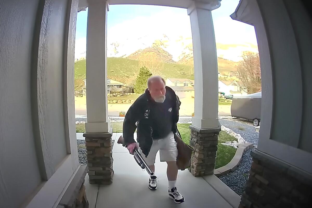 A man holding a shotgun and duffle bag is walking toward the camera and front door of a house. 