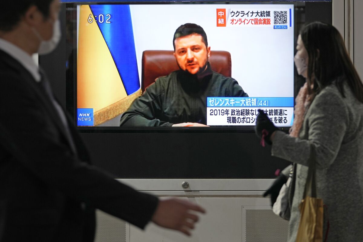 People walking past a TV screen showing a man seated next to a yellow and blue flag 