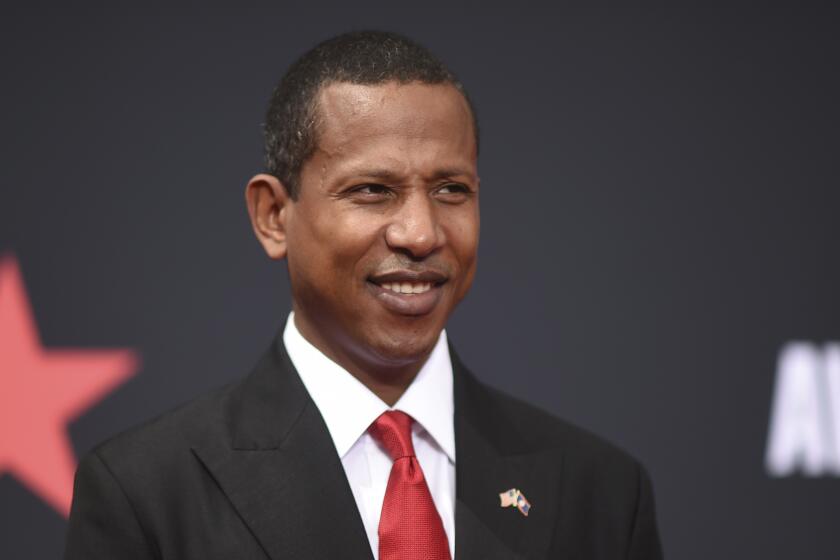 Shyne in a black suit and a red tie smiling and posing against a black background