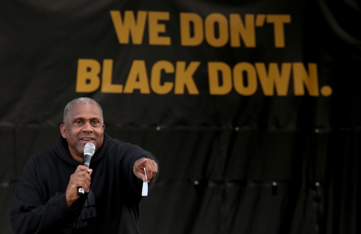 A man holds a microphone with  his right hand and points with the other in front of big words "We don't Black down."