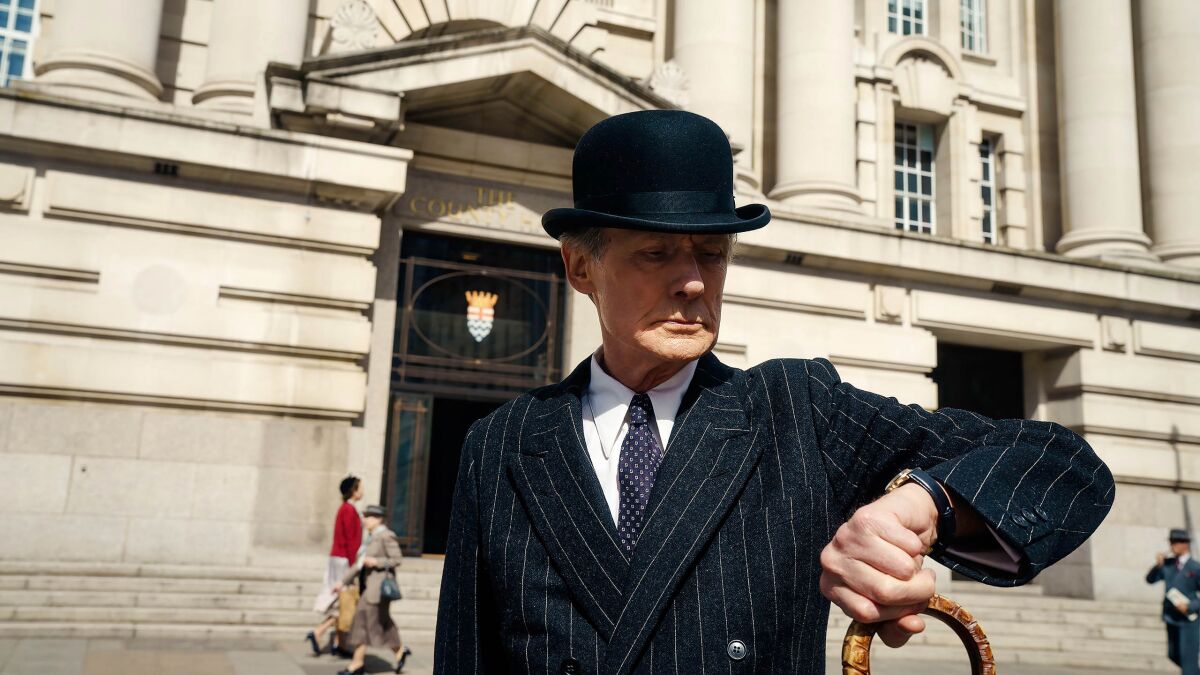 A man in a striped suit and bowler hat stands outside a large building and checks his watch in a scene from "Living."