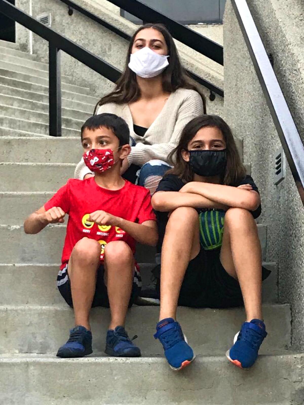 Three children wear masks and sit on steps.