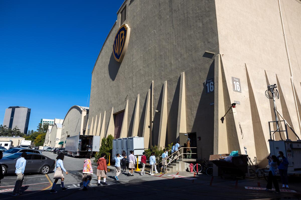 A single-file line of children walking into a large, beige building with the Warner Bros. logo on it.