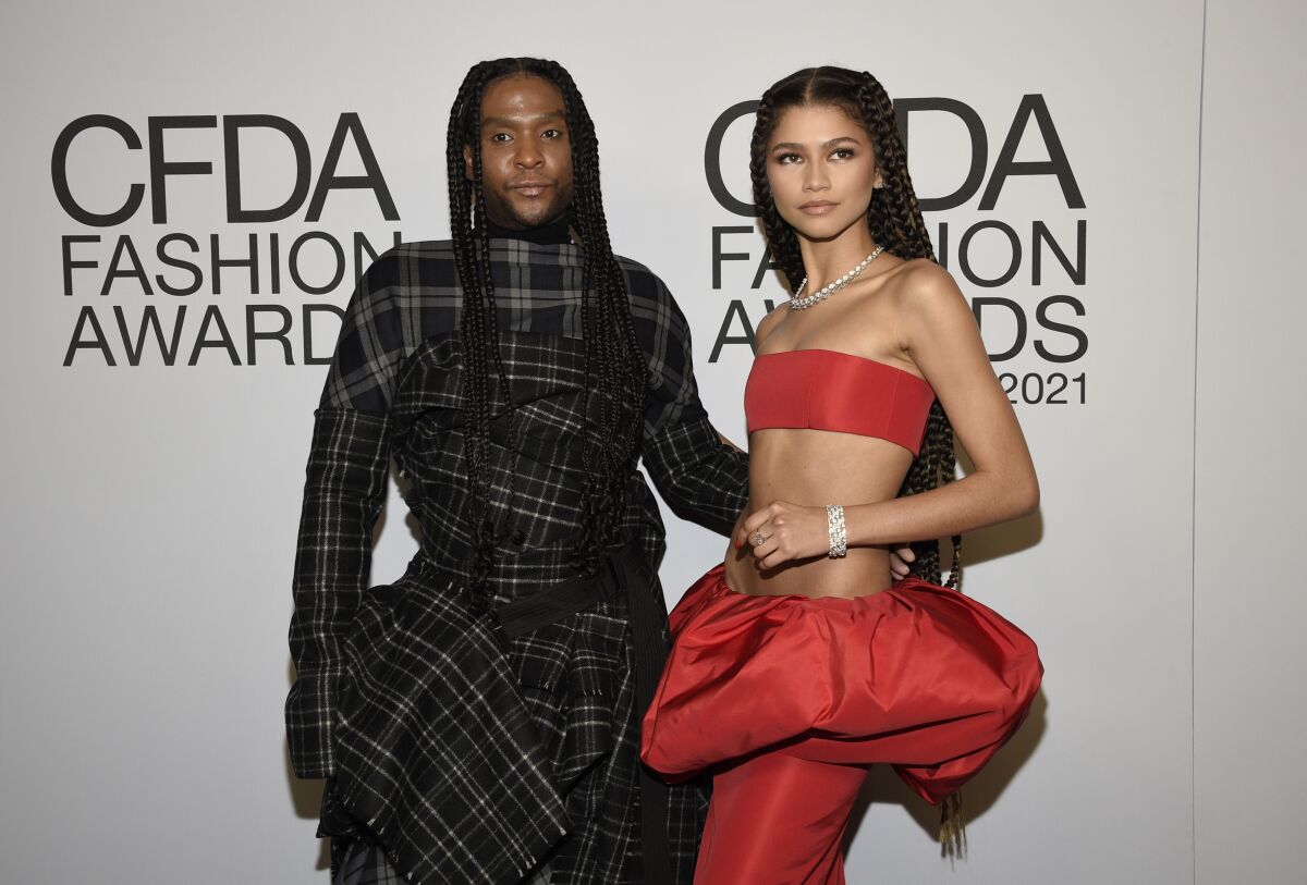 A man with braids in a dark plaid gown poses with his arm on the waist of a woman in a bright red bandeau and puffy skirt