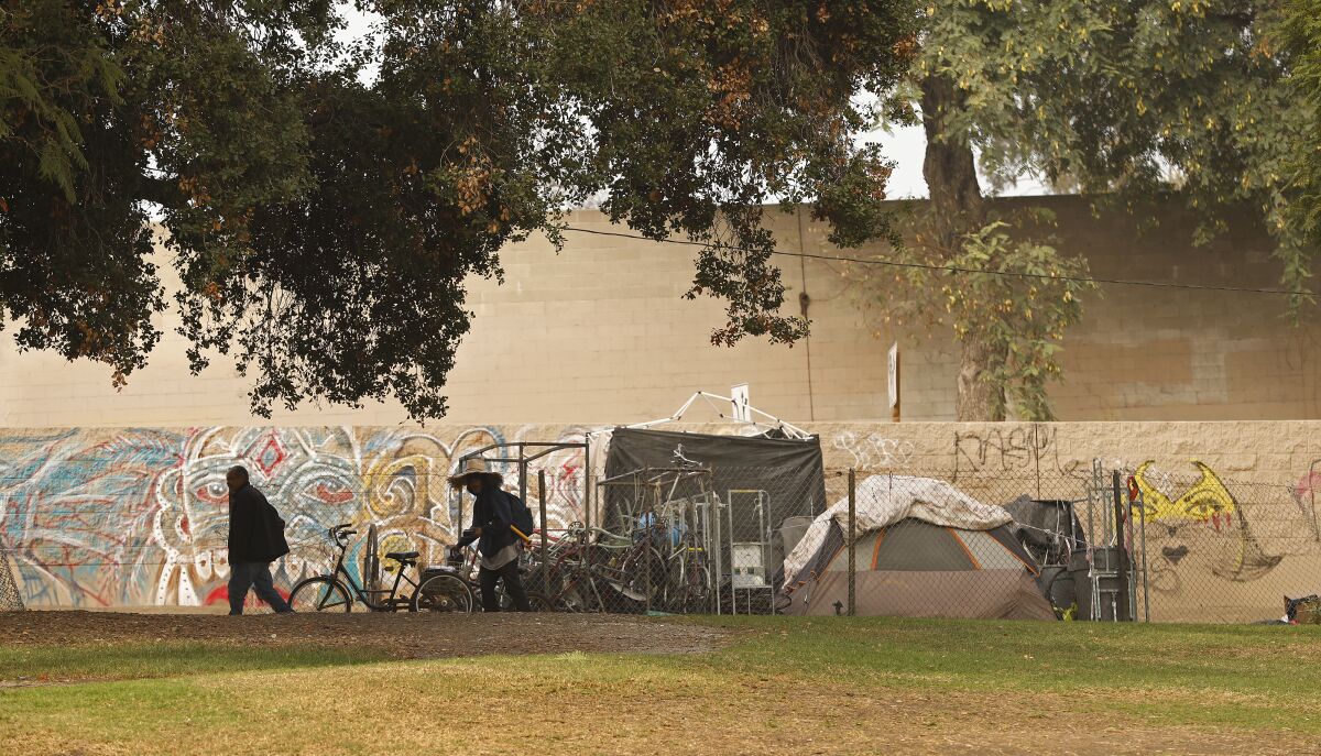 Two people walk past tents and bicycles clustered behind a chain-link fence at a park