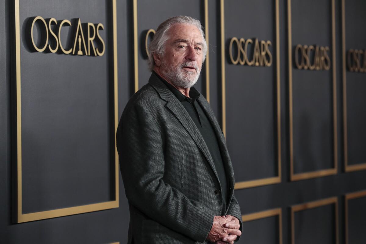 a man in a black shirt and jacket in front of a wall with the word "Oscars" repeated in gold 