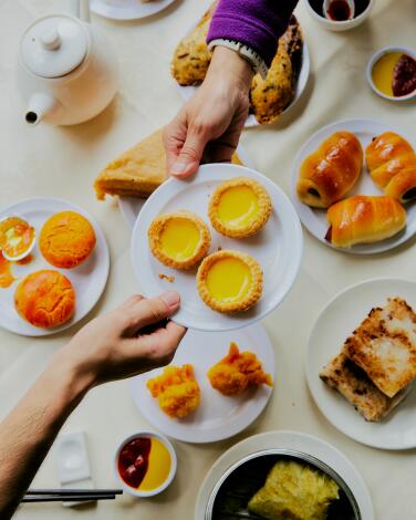 Hands pass a plate of egg tarts over a table laden with dim sum items