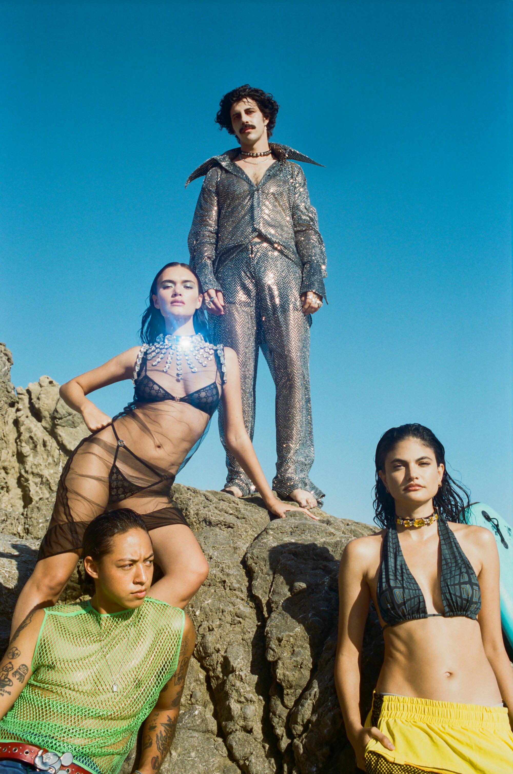 Four people wearing colorful, glittery outfits stand at the top and base of a rock on the beach.