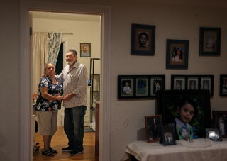 A couple standing in a bright room, framed by a doorway in an adjoining room with a wall covered in family photos.
