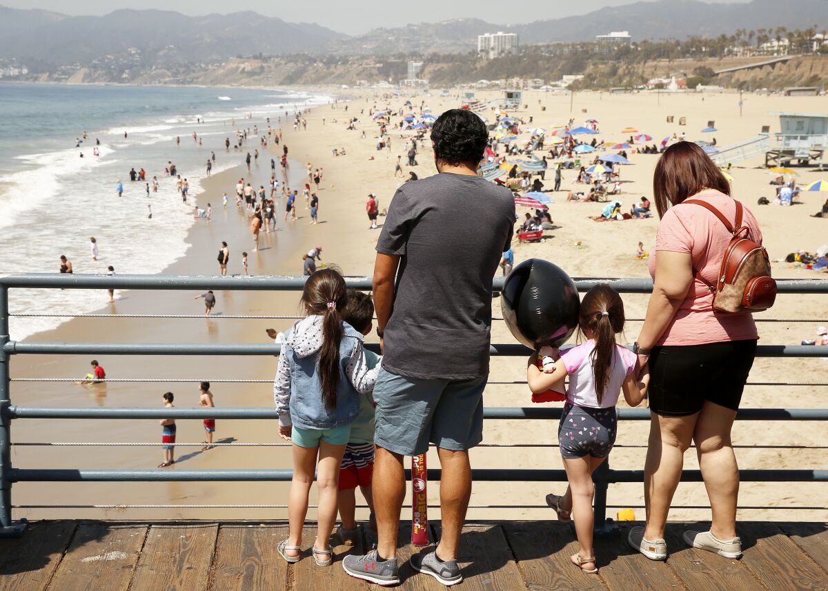A man and a woman stand with three young children at a pier railing overlooking beachgoers and the ocean. 