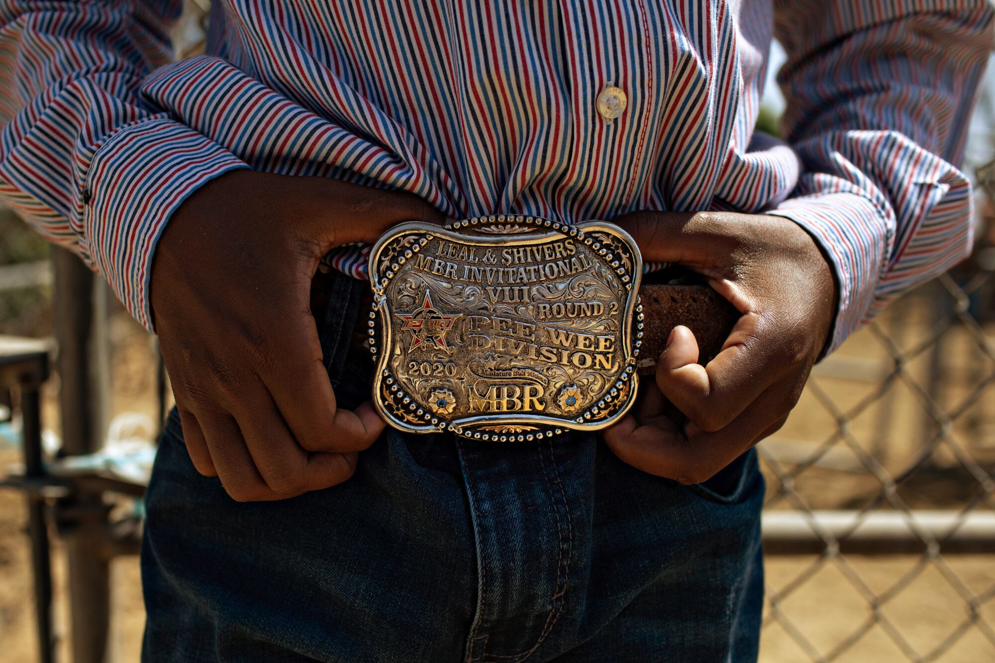 A closeup of a large rodeo championship belt buckle, worn by a boy in jeans and a striped shirt and framed by his hands