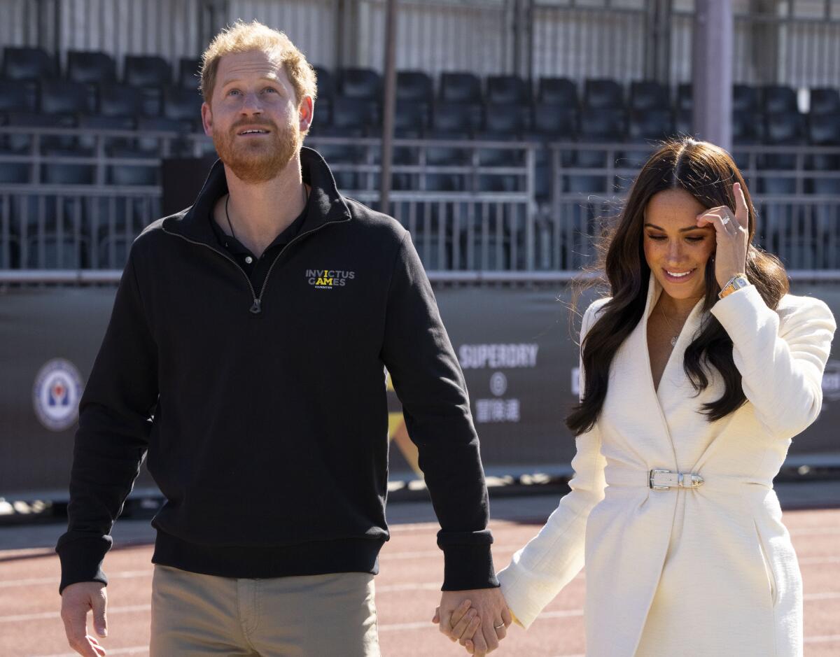 Harry and Meghan walk hand-in-hand on a track inside a stadium. Harry wears a black sweater; Meghan wears a white suit.