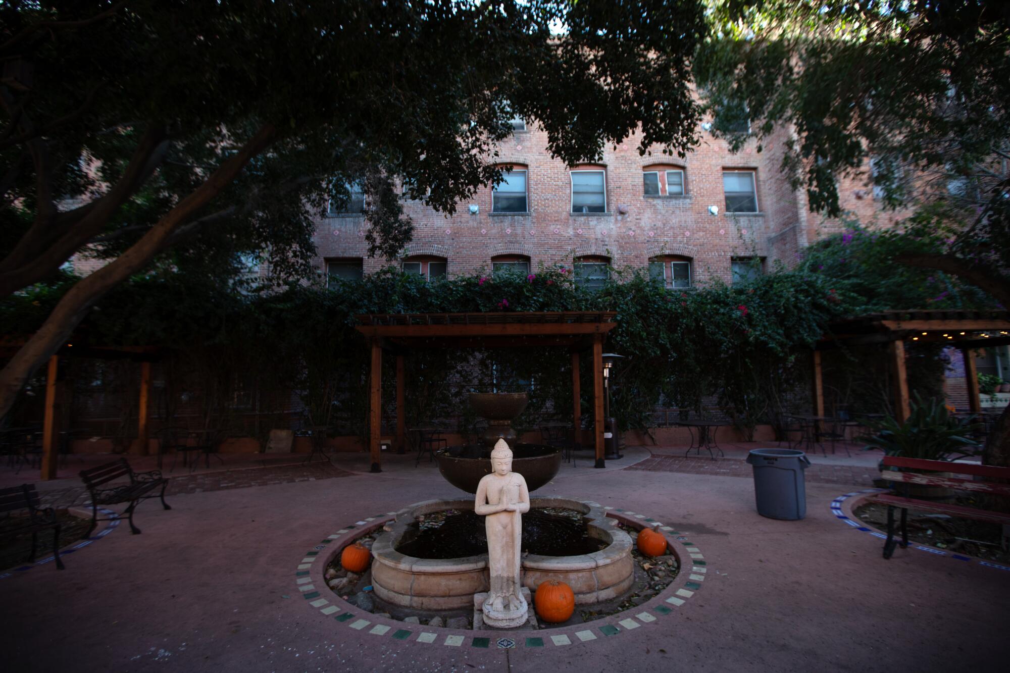 A small statue of a figure with folded hands stands near a dry fountain in a shaded courtyard.