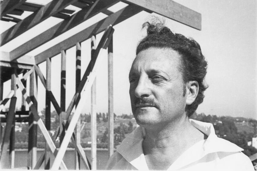 Rudolph Schindler, sporting a white shirt open at the neck, his hair in a pompadour, stands before the frame of a house.