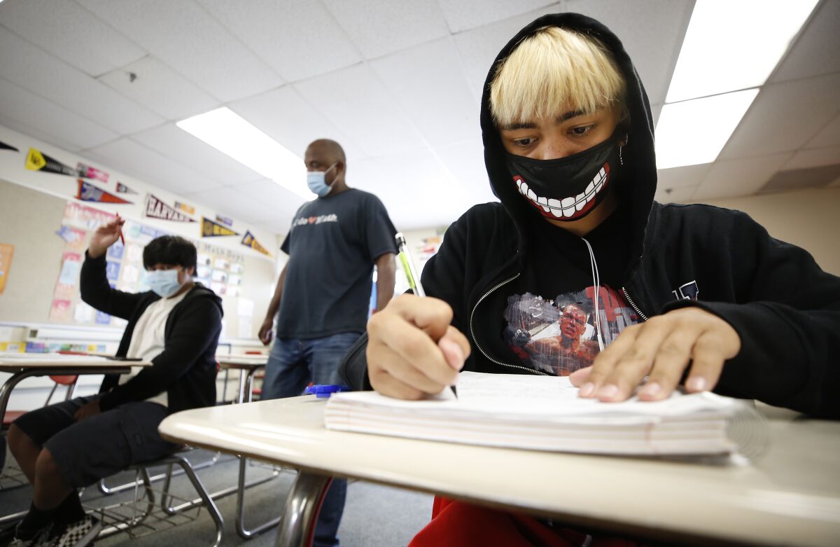 A high school boy at his desk wears a mask imprinted with a smiling mouth.