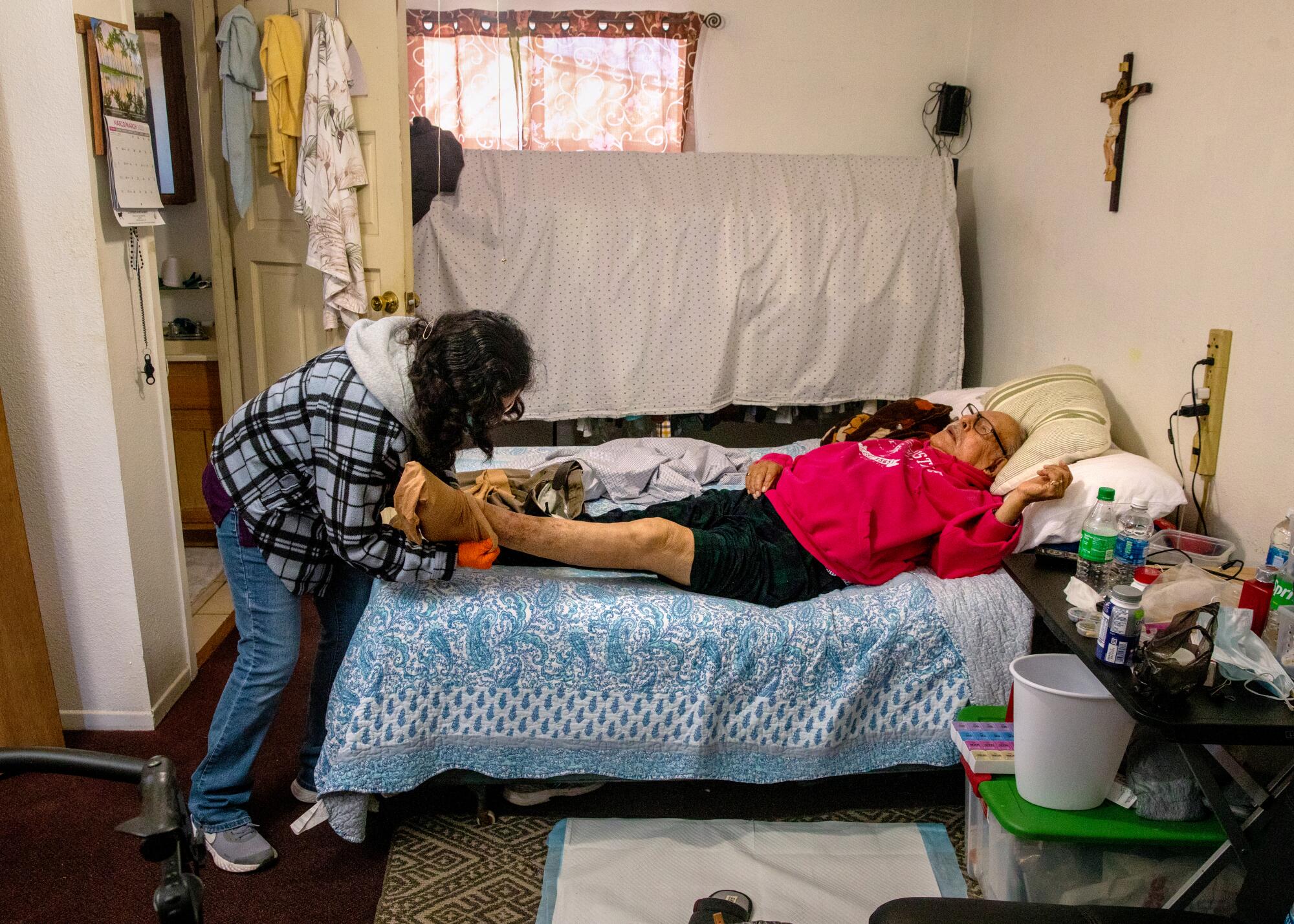 A woman puts compression stockings on an elderly man lying on a bed.