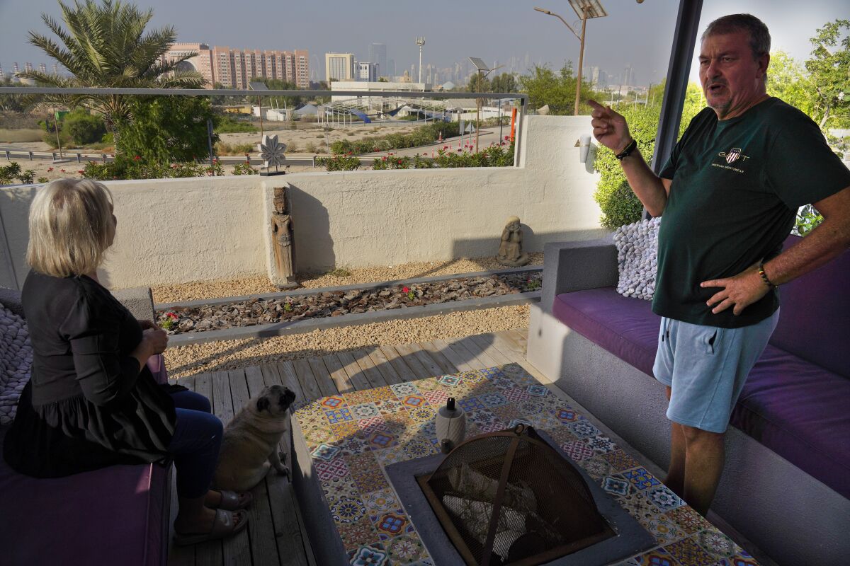 Garry James, right, speaks to The Associated Press as his wife Amanda James and their pug Bella sit out on their front porch overlooking what once was empty desert in Jebel Ali Village in Dubai, United Arab Emirates, Tuesday, Nov. 2, 2021. Nakheel, the state-owned developer of Dubai's signature palm-shaped islands, has unveiled plans to demolish the neighborhood to make way for a gated community of flashy, two-story villas. Residents of the village, which dates back to the late 1970s, have received eviction notices and say they're upset to leave the quiet neighborhood. (AP Photo/Jon Gambrell)
