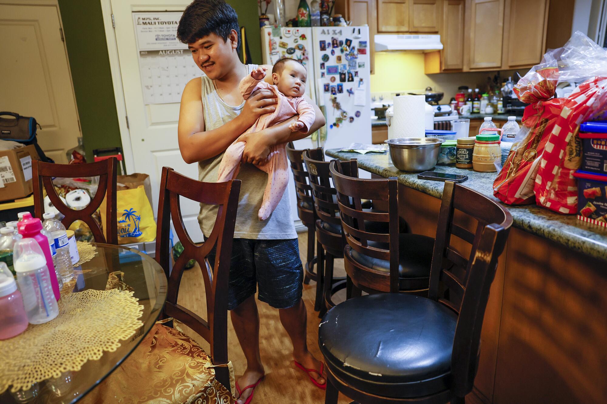 A man in a sleeveless shirt and shorts holds a baby in a pink outfit while standing near a dining table and chairs 
